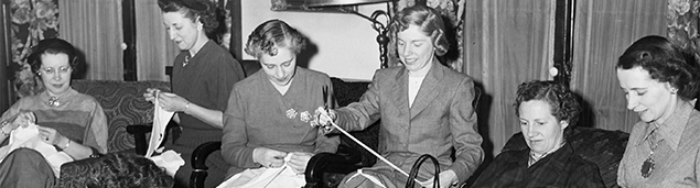 a black and white photograph of six women sitting in a room sewing together