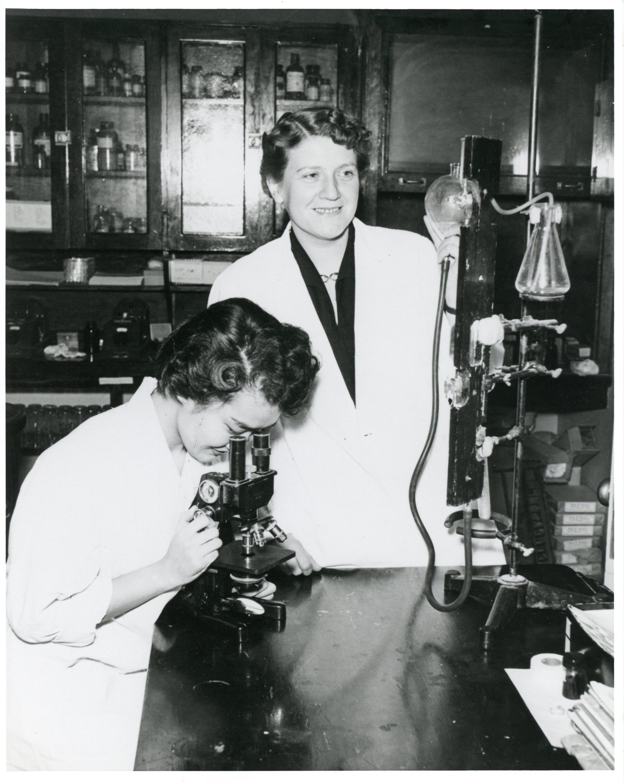 black and white photo of a woman looking in a microscope while another woman looks on
