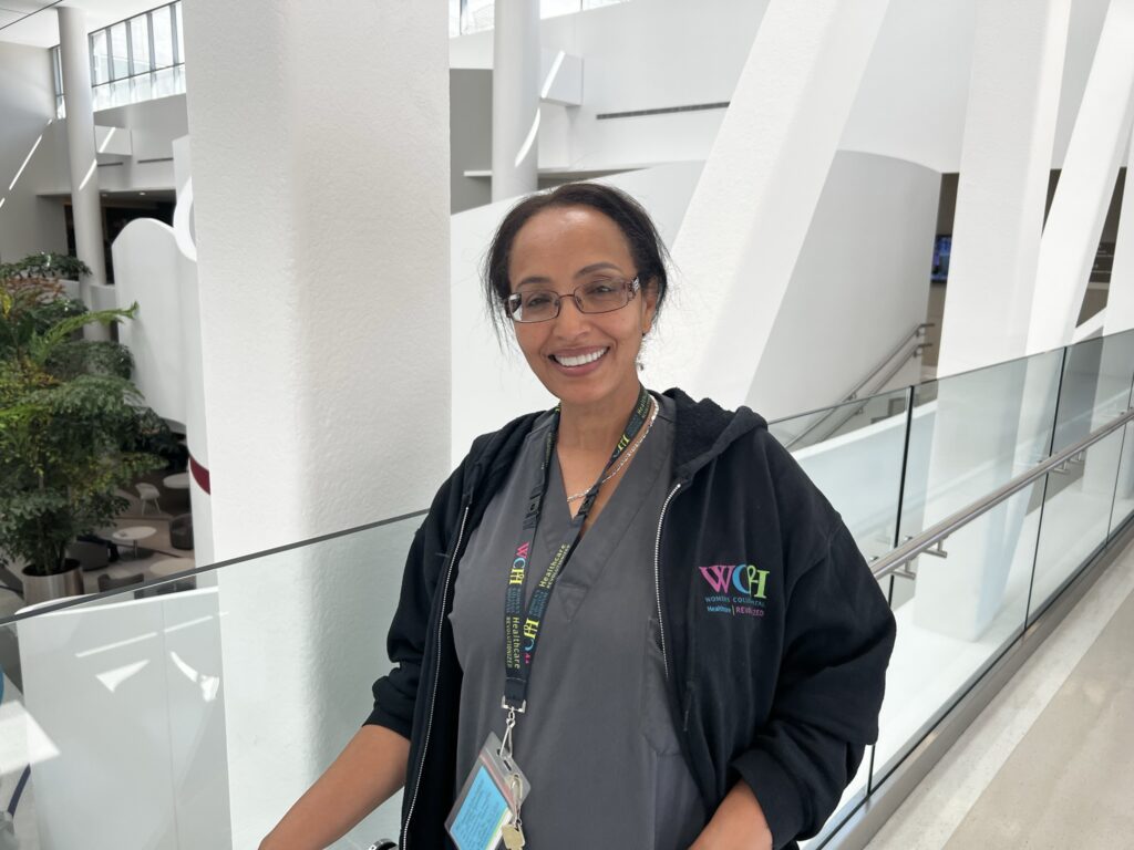 WCH EVS worker Asmakch Ayeneababa, photographed on the bridge at the hospital wearing grey scrubs and a black WCH-branded hoodie